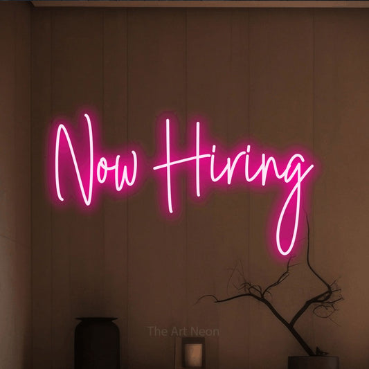 Hot pink script-style LED neon sign reading "Now Hiring" against a warm brown wall for restaurant décor