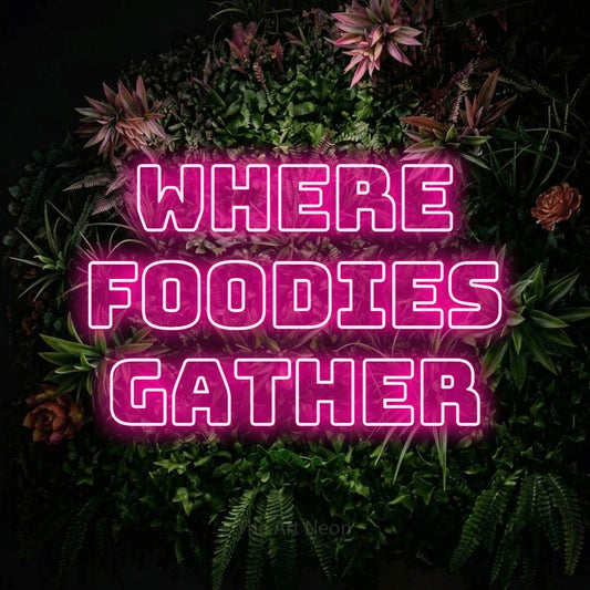 Hot pink outline-style LED neon sign reading "Where Foodies Gather" against a flowering garden wall for restaurant décor