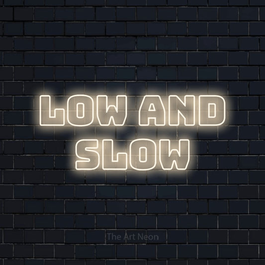 Warm white outline-style LED neon sign reading "Low and Slow" against a dark navy brick wall for restaurant décor