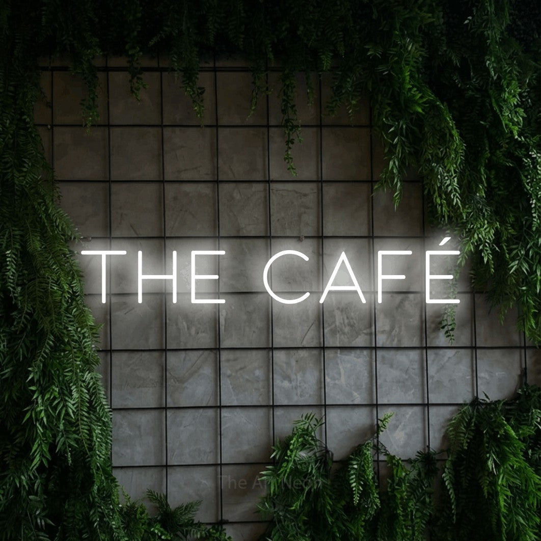 Cool white LED neon sign reading "The Café" against a green grid wall with foliage for café décor