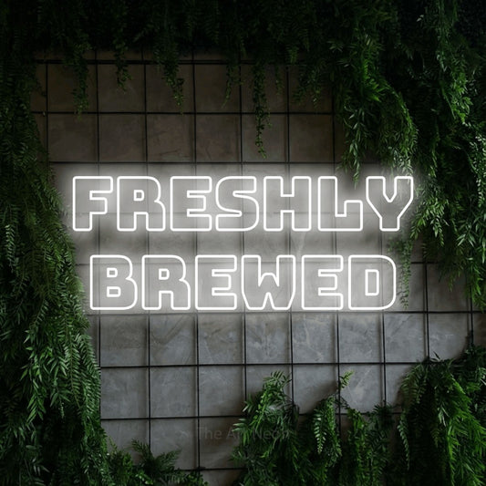 Cool white outline-style LED neon sign reading "Freshly Brewed" against a green grid wall with foliage for café décor