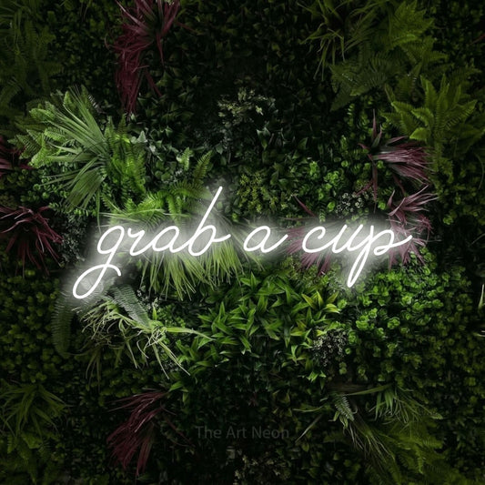 Cool white script-style LED neon sign reading "Grab a Cup" against a lush garden wall with pink flowers for café décor