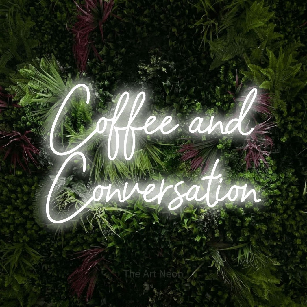 Cool white script-style LED neon sign reading "Coffee and Conversation" against a lush garden wall with pink flowers