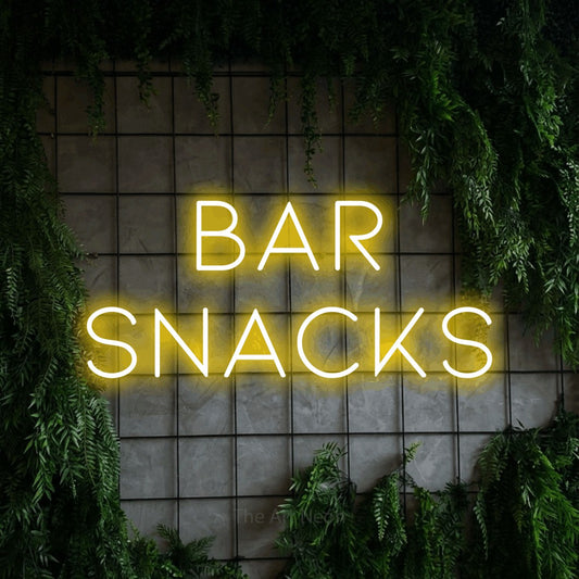 Yellow LED neon sign reading "Bar Snacks" against a green grid wall with foliage for bar décor