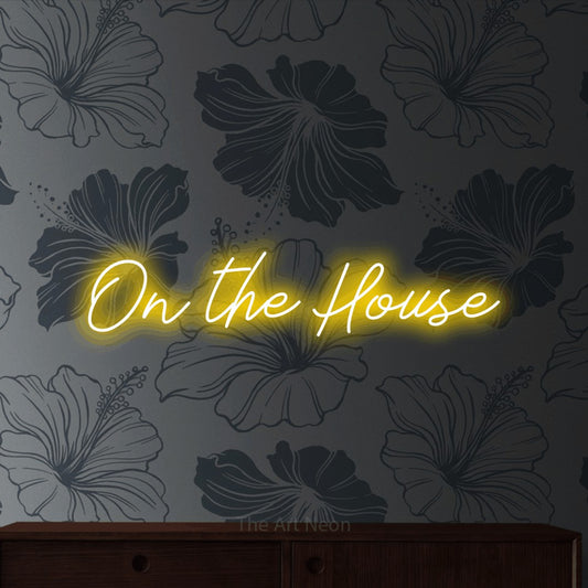 Yellow script-style LED neon sign reading "On the House" against grey floral wallpaper for bar décor