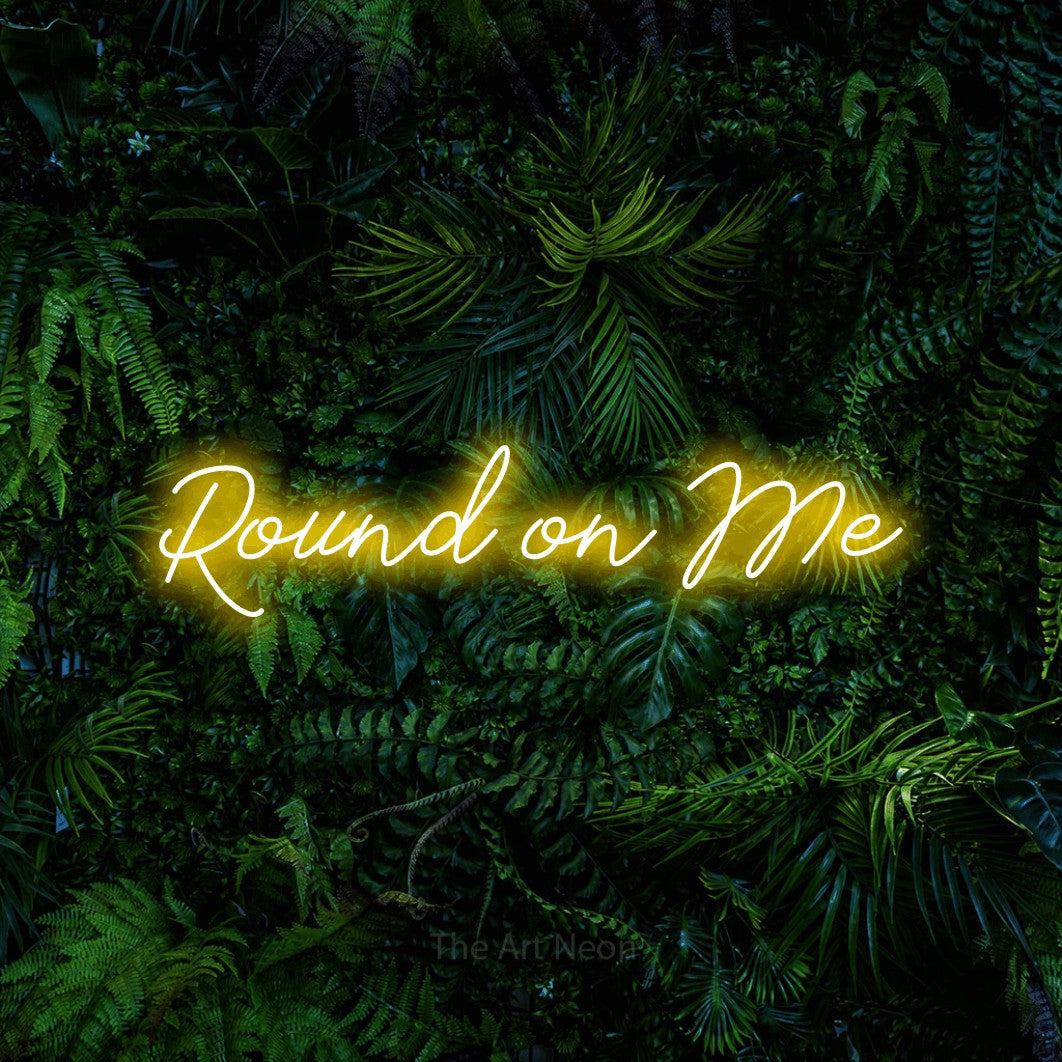 Yellow script-style LED neon sign reading "Round on Me" against a dense tropical foliage wall for bar décor