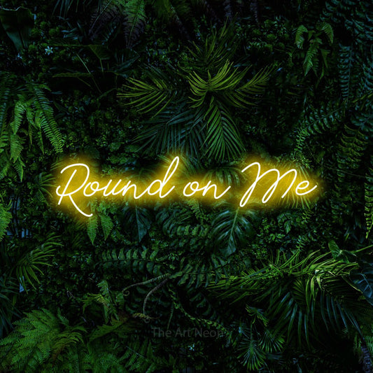 Yellow script-style LED neon sign reading "Round on Me" against a dense tropical foliage wall for bar décor