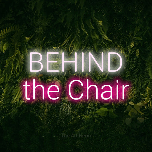 Hot pink and cool white LED neon sign reading "Behind the Chair" against a moss-covered green wall for salon décor