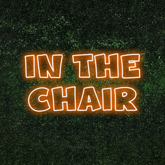 Orange outline-style LED neon sign reading "In the Chair" against a textured green moss wall for salon décor