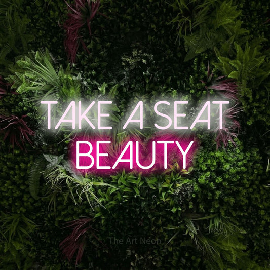 Hot pink and cool white LED neon sign reading "Take a Seat Beauty" against a lush garden wall with pink flowers