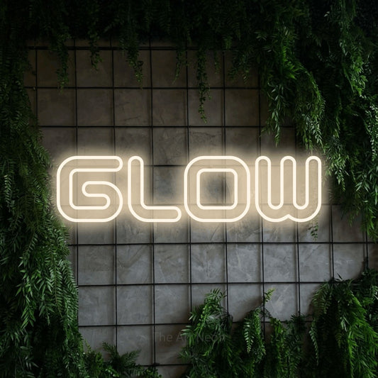 Warm white outline-style LED neon sign reading "Glow" against a green grid wall with foliage for salon décor
