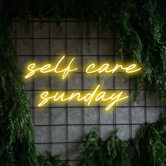 Yellow LED neon sign reading "Self Care Sunday" against a green grid wall with foliage for salon décor