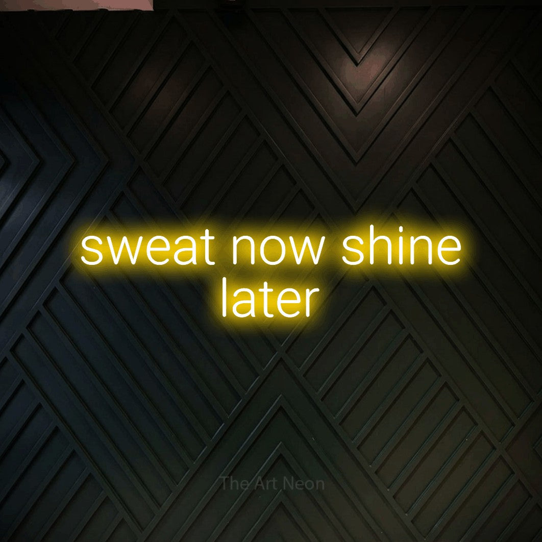 Yellow LED neon sign reading "Sweat Now Shine Later" against a dark geometric wood wall for gym décor