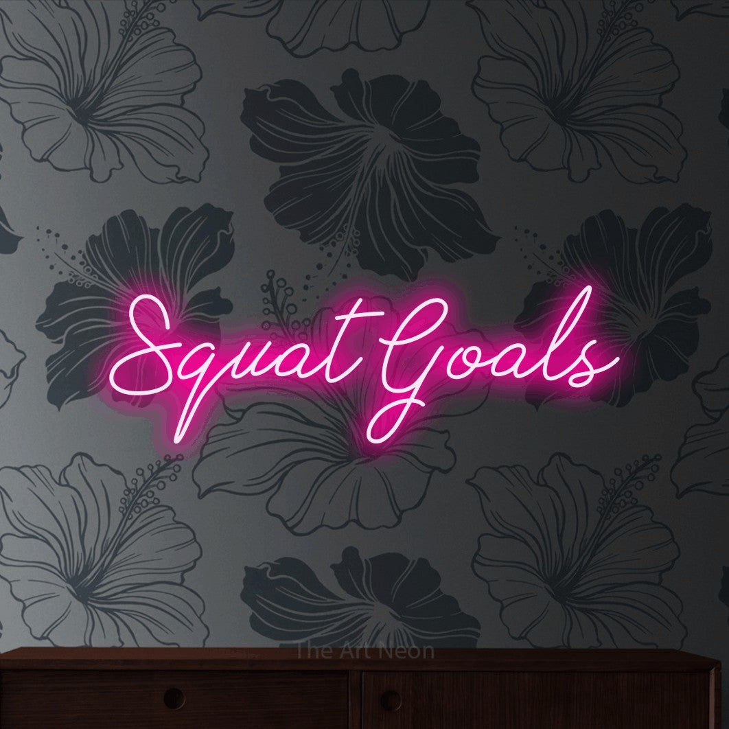 Hot pink script-style LED neon sign reading "Squat Goals" against grey floral wallpaper for gym décor