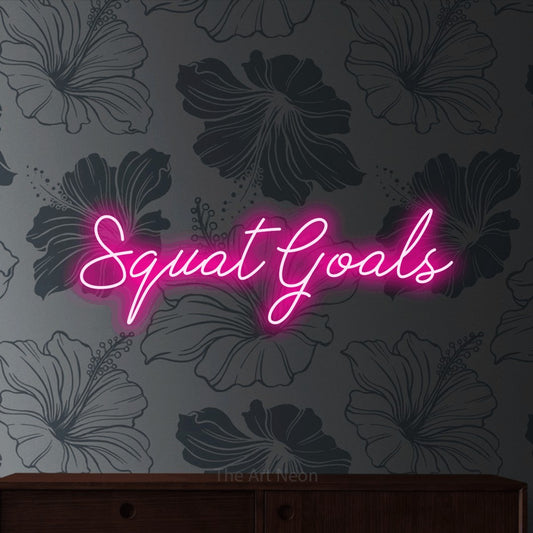Hot pink script-style LED neon sign reading "Squat Goals" against grey floral wallpaper for gym décor