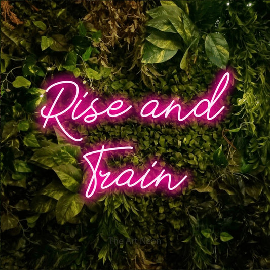 Hot pink script-style LED neon sign reading "Rise and Train" against a textured moss and foliage wall for gym décor