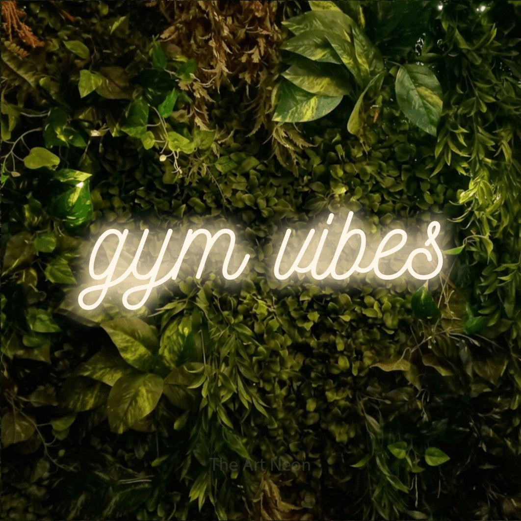 Enseigne néon à DEL à lumière blanche chaude, de style manuscrit, affichant « Gym Vibes » sur un mur texturé de mousse et de feuillage, pour décorer une salle d’entraînement