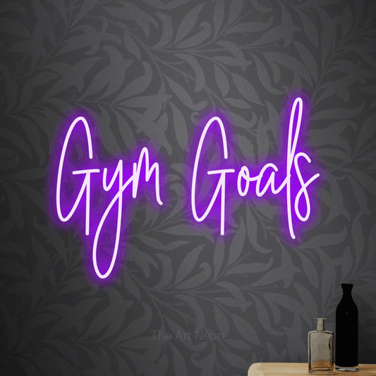 Purple script-style LED neon sign reading "Gym Goals" against light grey floral wallpaper for gym décor