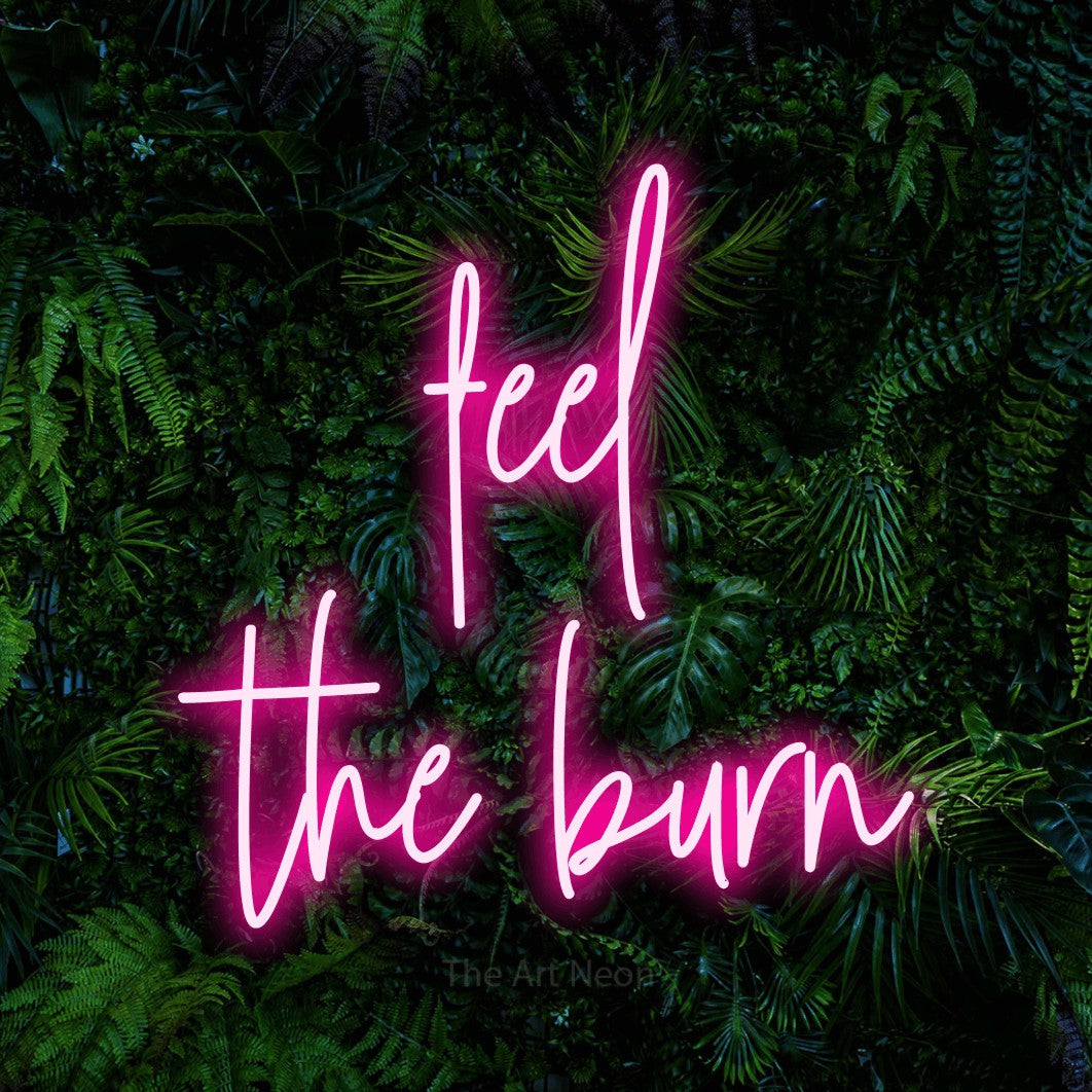 Hot pink script-style LED neon sign reading "Feel the Burn" against a dense tropical foliage wall for gym décor