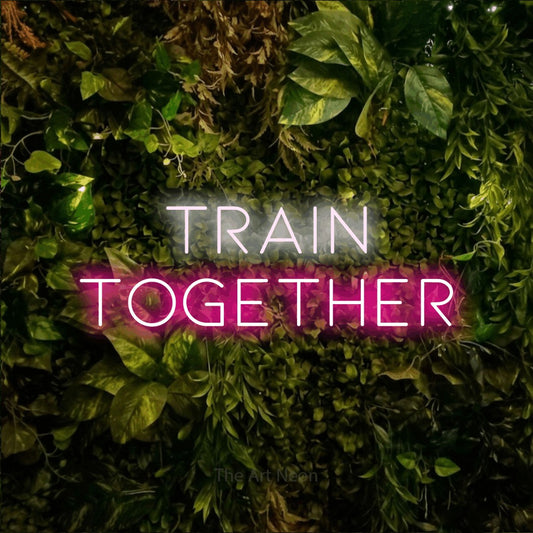 Hot pink and cool white LED neon sign reading "Train Together" against a textured moss and foliage wall for gym décor