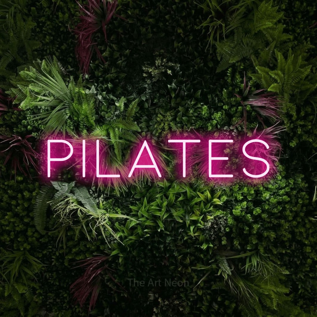 Enseigne néon à DEL à lumière rose vif, affichant « Pilates » sur un mur de jardin luxuriant aux fleurs roses, pour décorer une salle d’entraînement