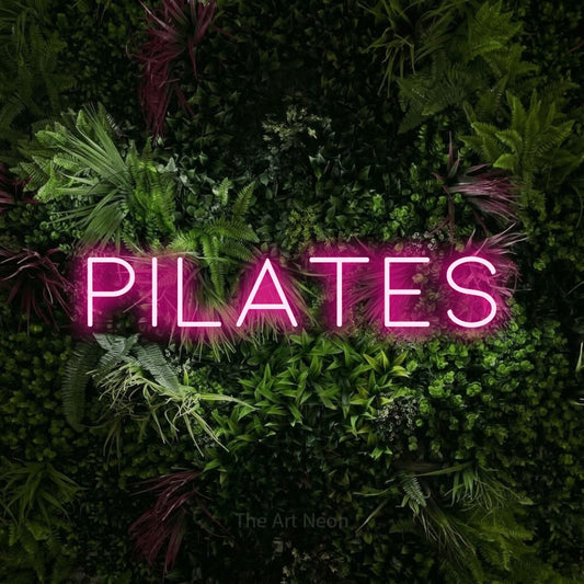 Hot pink LED neon sign reading "Pilates" against a lush garden wall with pink flowers for gym décor