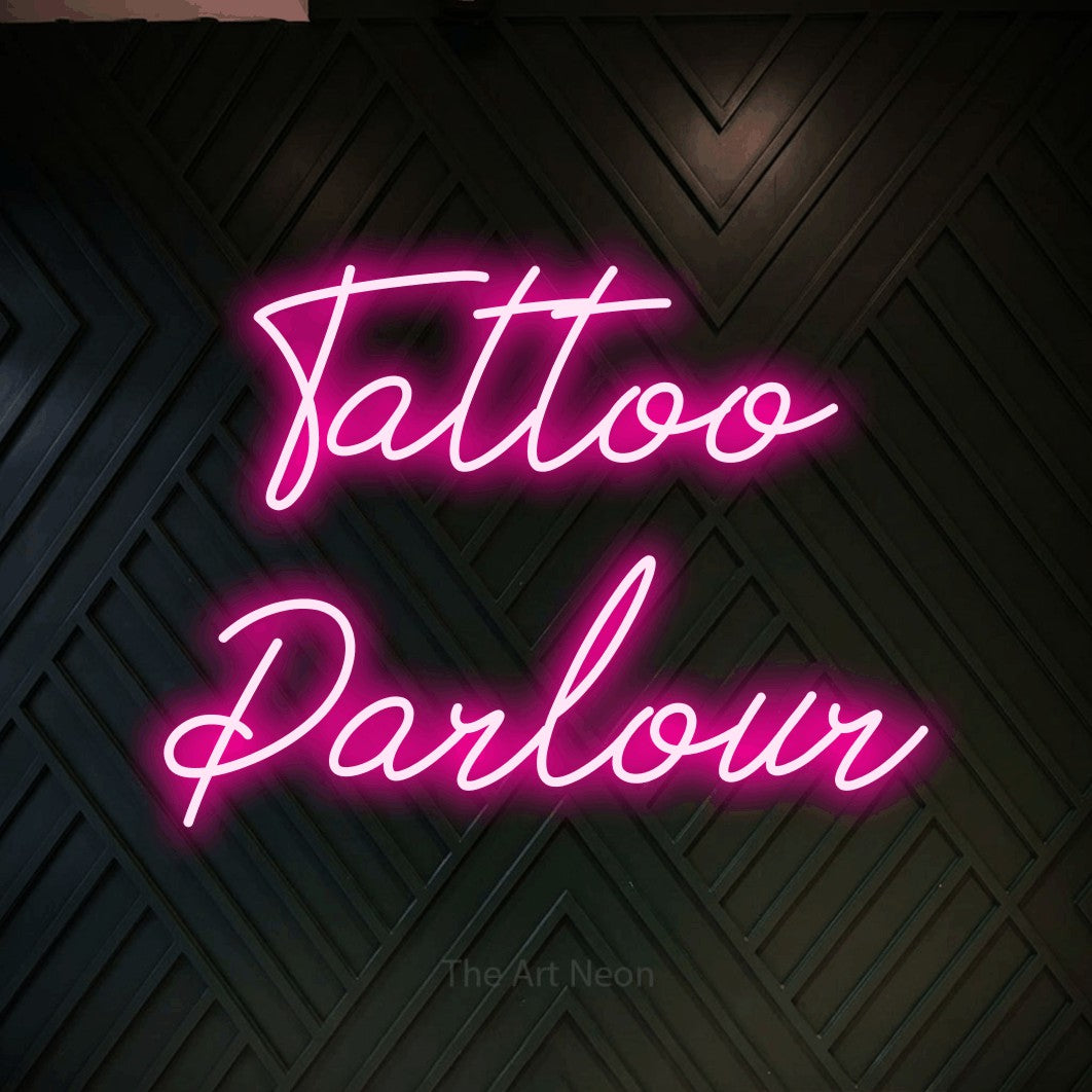 Hot pink script-style LED neon sign reading "Tattoo Parlour" against a dark geometric wood wall for tattoo studio décor