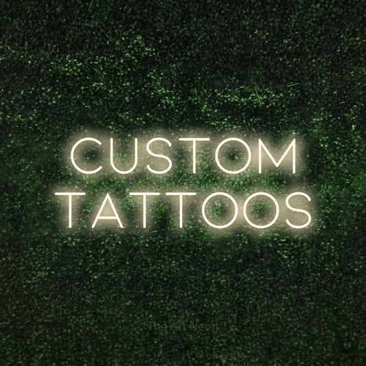 Warm white LED neon sign reading "Custom Tattoos" against a textured green moss wall for tattoo studio décor