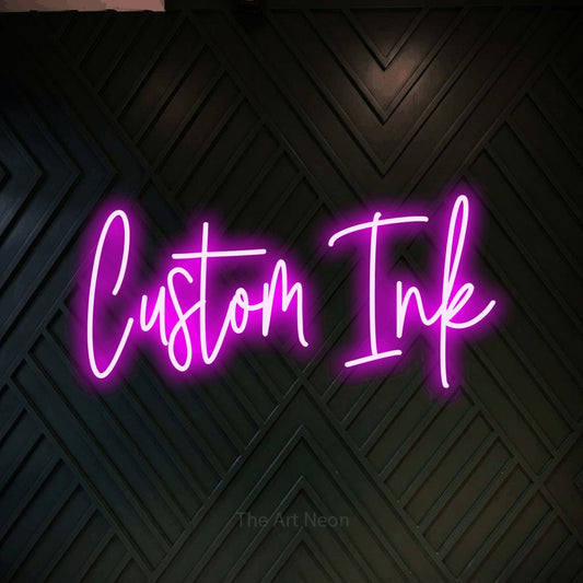 Pink script-style LED neon sign reading "Custom Ink" against a dark geometric wood wall for tattoo studio décor