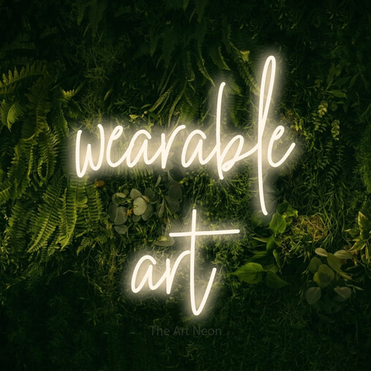 Warm white script-style LED neon sign reading "Wearable Art" against a moss-covered green wall for tattoo studio décor