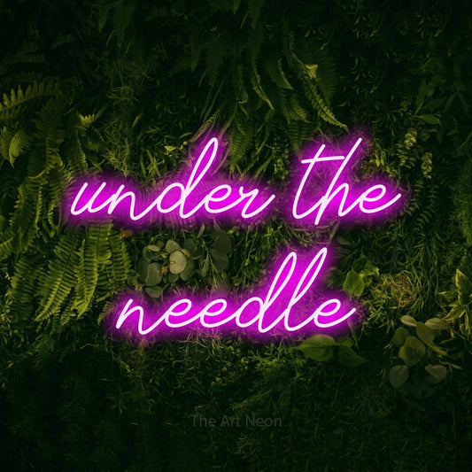 Pink script-style LED neon sign reading "Under the Needle" against a moss-covered green wall for tattoo studio décor