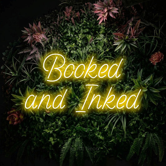 Yellow LED neon sign reading "Booked and Inked" against a flowering garden wall for tattoo studio décor
