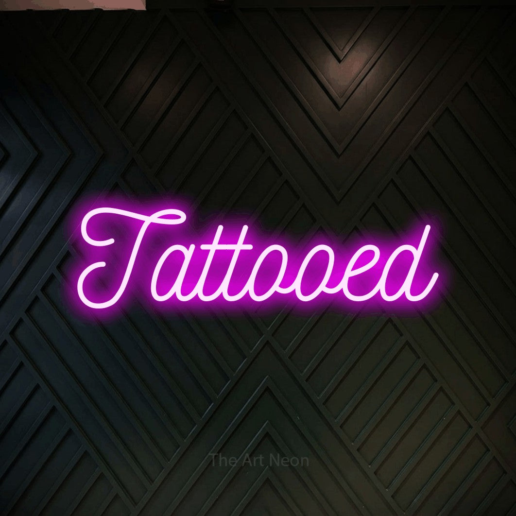 Pink script-style LED neon sign reading "Tattooed" against a dark geometric wood wall for tattoo studio décor