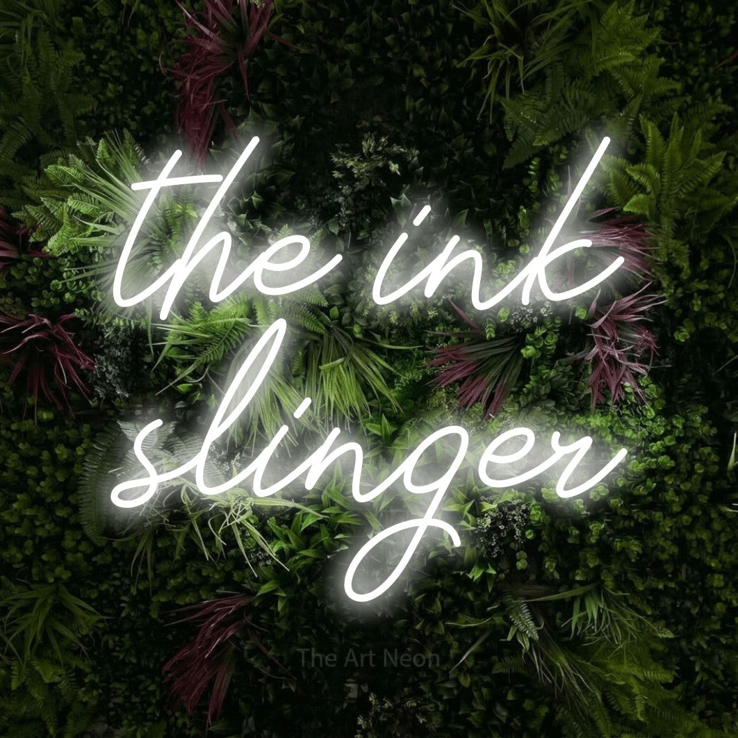 Cool white script-style LED neon sign reading "The Ink Slinger" against a lush garden wall with pink flowers
