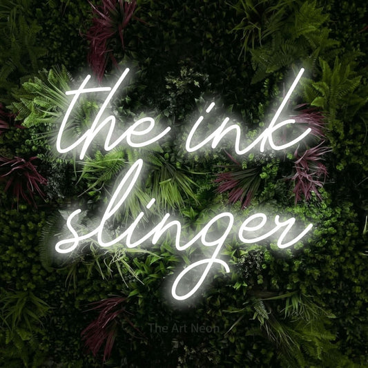 Cool white script-style LED neon sign reading "The Ink Slinger" against a lush garden wall with pink flowers