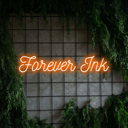 Orange script-style LED neon sign reading "Forever Ink" against a green grid wall with foliage for tattoo studio décor