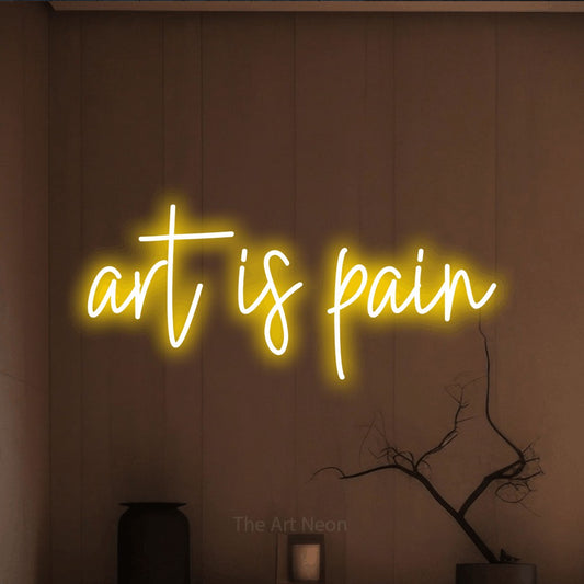 Yellow script-style LED neon sign reading "Art is Pain" against a warm brown wall for tattoo studio décor