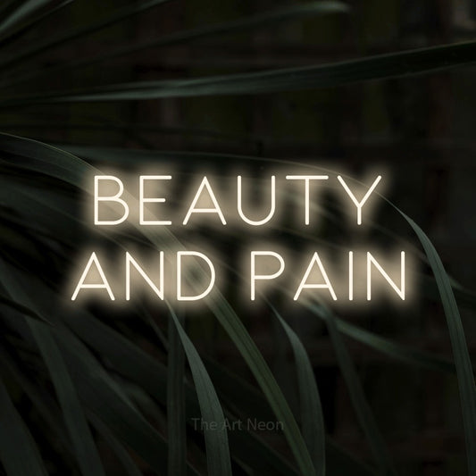 Warm white LED neon sign reading "Beauty and Pain" against dark tropical leaves for tattoo studio décor