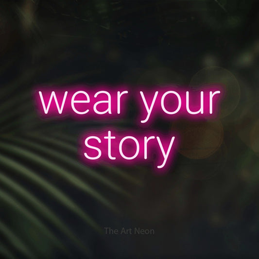 Hot pink LED neon sign reading "Wear Your Story" against dark blurred palm leaves for tattoo studio décor