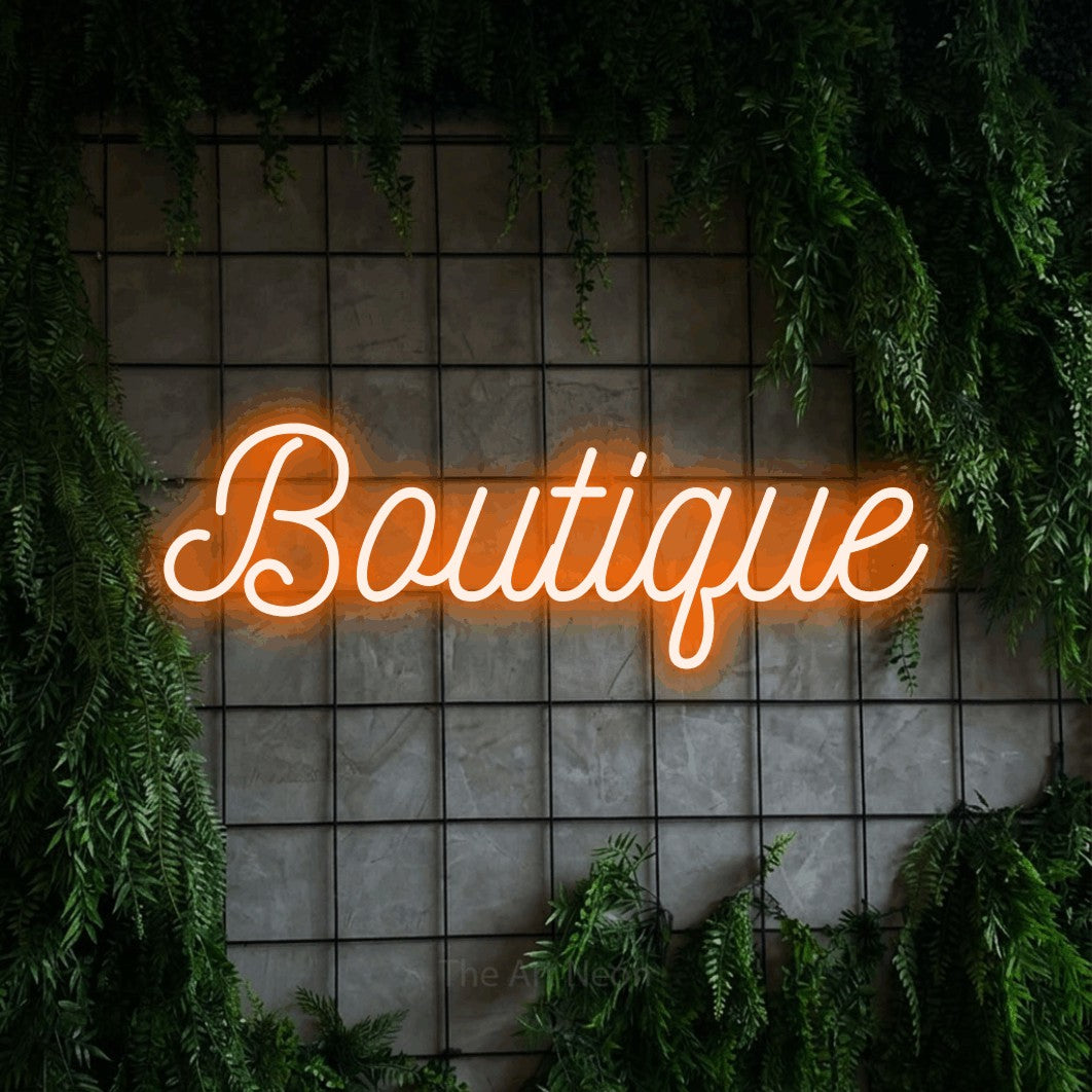Orange script-style LED neon sign reading "Boutique" against a green grid wall with foliage
