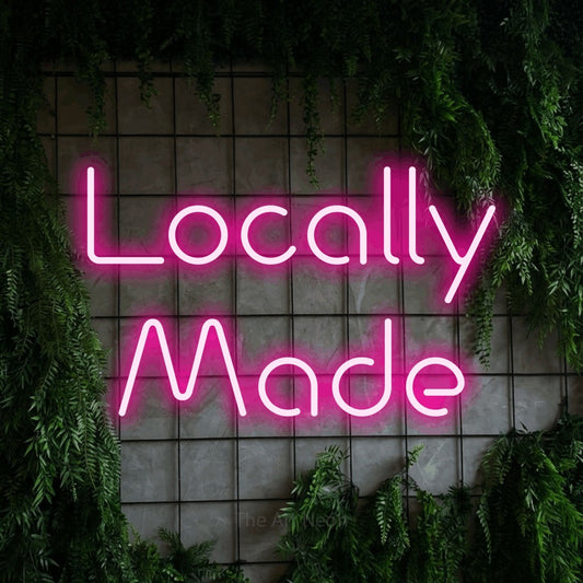 Hot pink LED neon sign reading "Locally Made" against a green grid wall with foliage