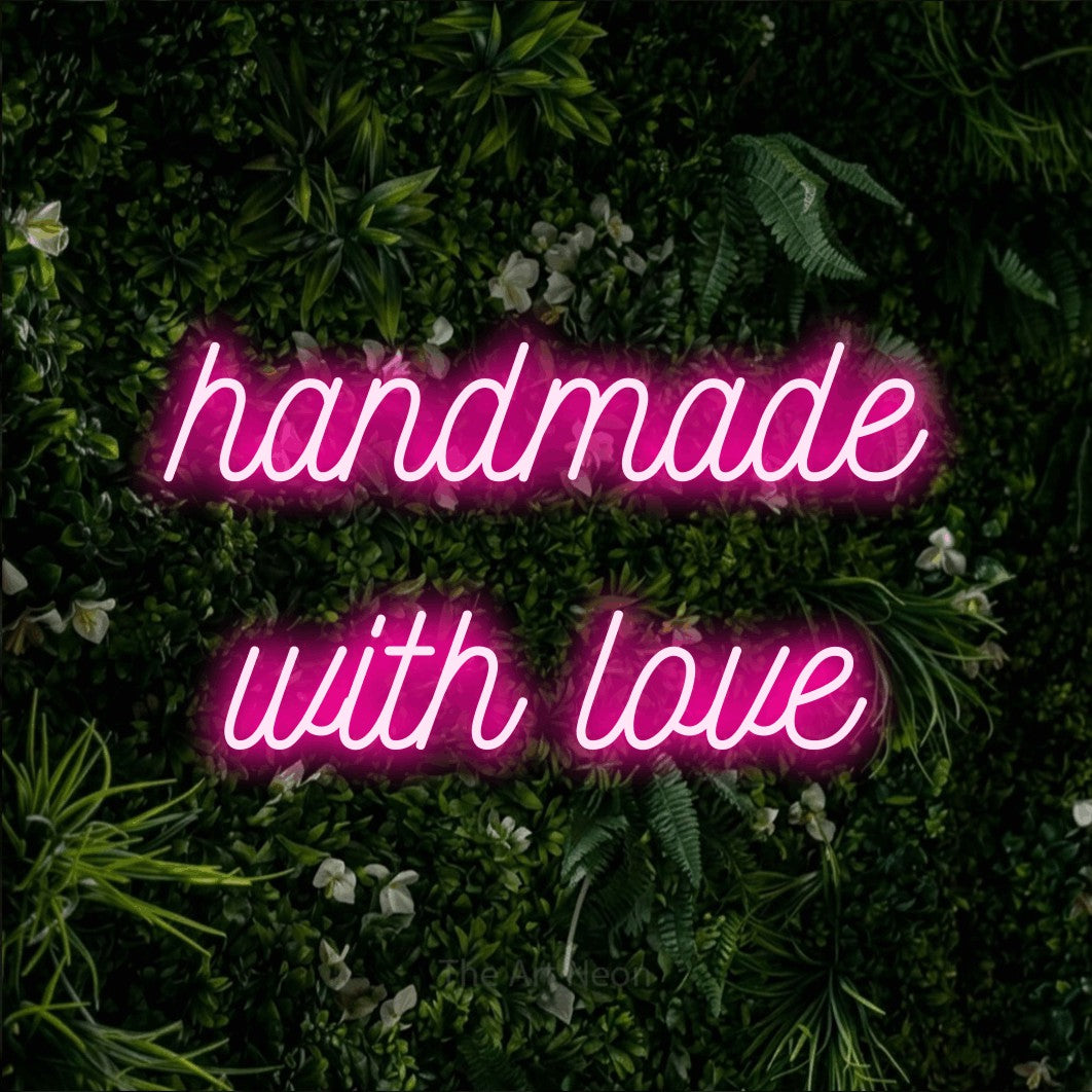 Hot pink script-style LED neon sign reading "Handmade with Love" against a leafy green wall