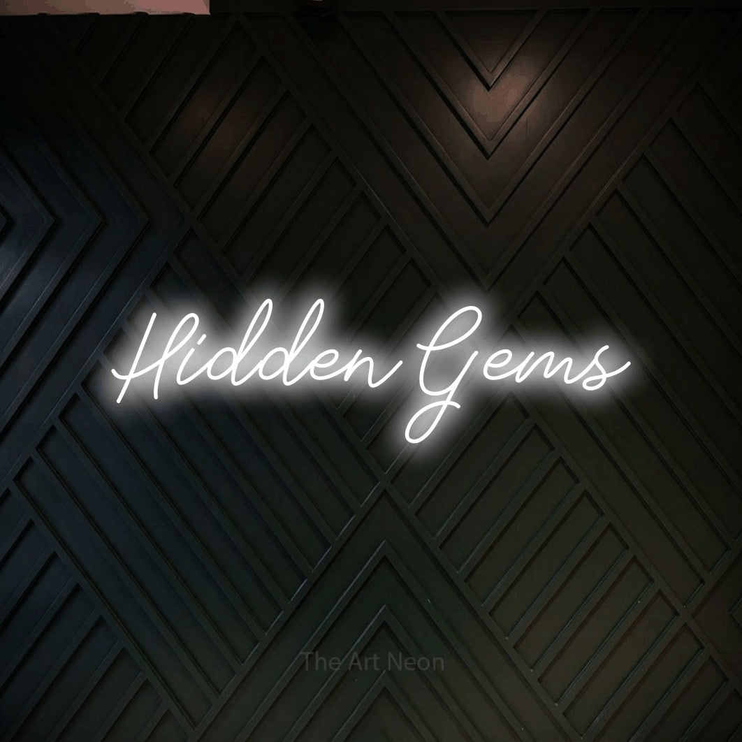 Cool white script-style LED neon sign reading "Hidden Gems" against a dark geometric wood wall