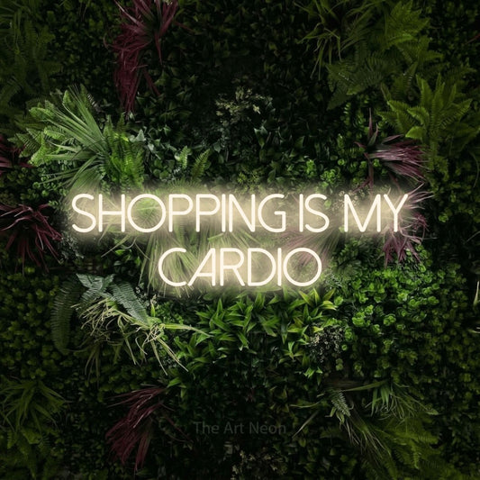 Warm white LED neon sign reading "Shopping is My Cardio" against a lush garden wall with pink flowers