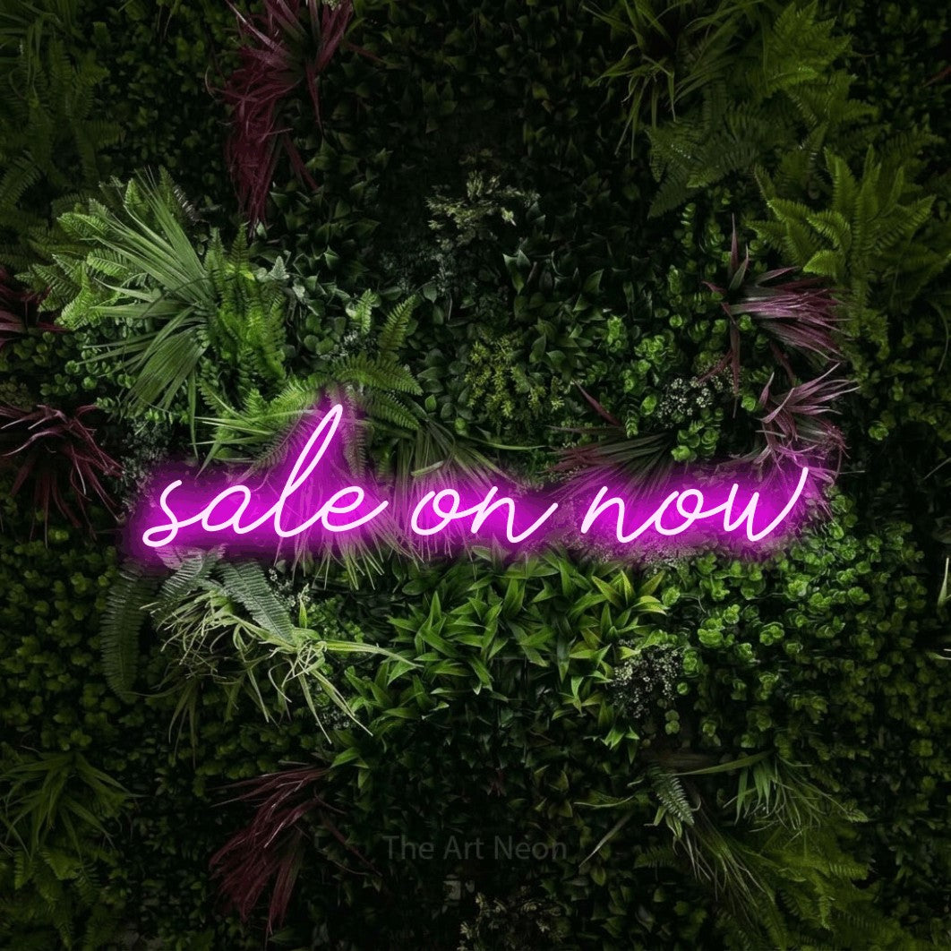 Pink script-style LED neon sign reading "Sale On Now" against a lush garden wall with pink flowers