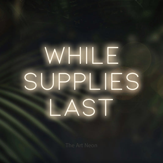 Warm white LED neon sign reading "While Supplies Last" against dark blurred palm leaves