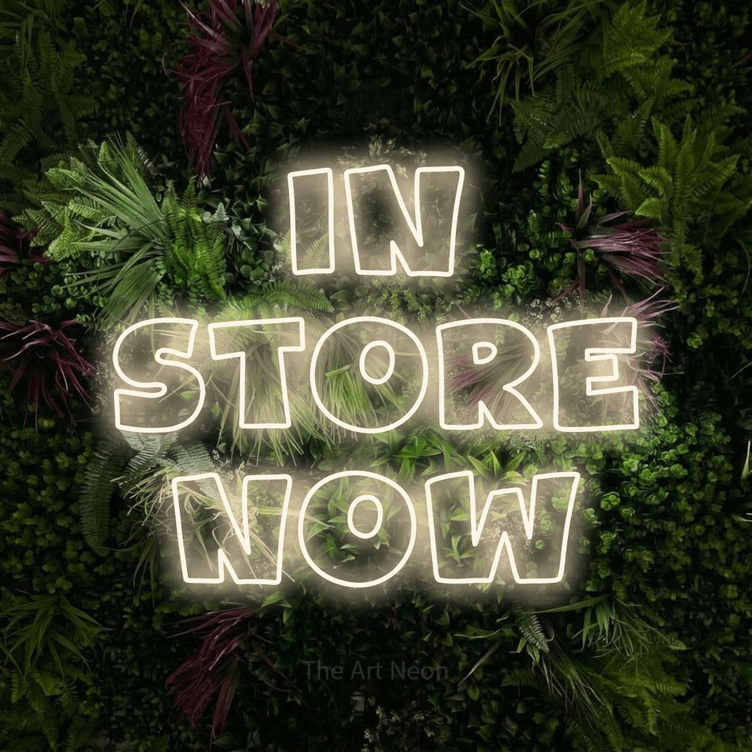 Warm white outline-style LED neon sign reading "In Store Now" against a lush garden wall with pink flowers