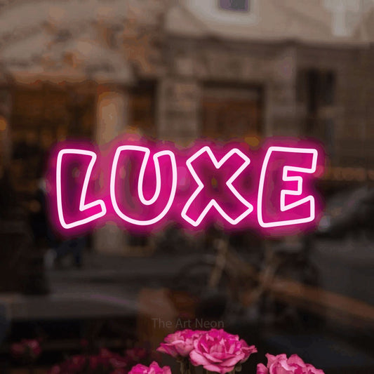 Hot pink outline-style LED neon sign reading "Luxe" in a softly blurred café interior