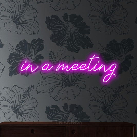 Pink script-style LED neon sign reading "In a Meeting" against grey floral wallpaper for office décor