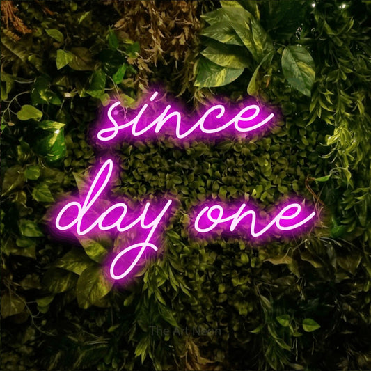 Pink script-style LED neon sign reading "Since Day One" against a textured moss and foliage wall for office décor